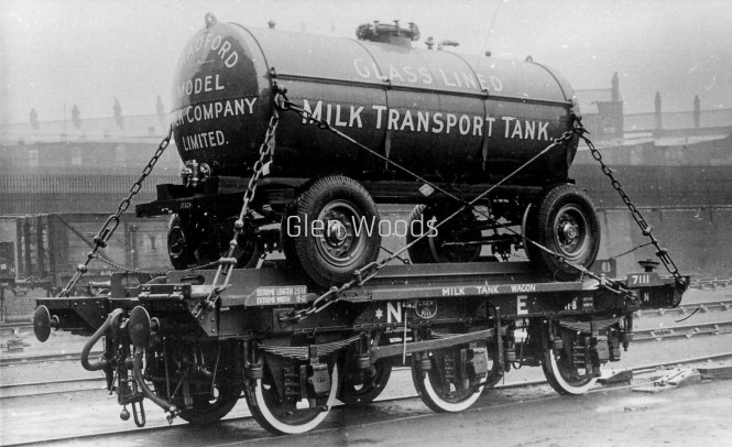 A black-and-white historical photograph of a specialized railway wagon carrying a large road tanker trailer on a set of tracks. The large cylindrical tank is painted a dark color with white lettering that reads "GLASS LINED MILK TRANSPORT TANK" on the side, and "BRADFORD MODEL MILK COMPANY LIMITED" on the circular end. The tanker trailer has large rubber tires and is securely chained down to a flatbed railway carriage labeled "MILK TANK WAGON" and marked with "NE" branding. A prominent "Glen Woods" watermark is centered across the image.