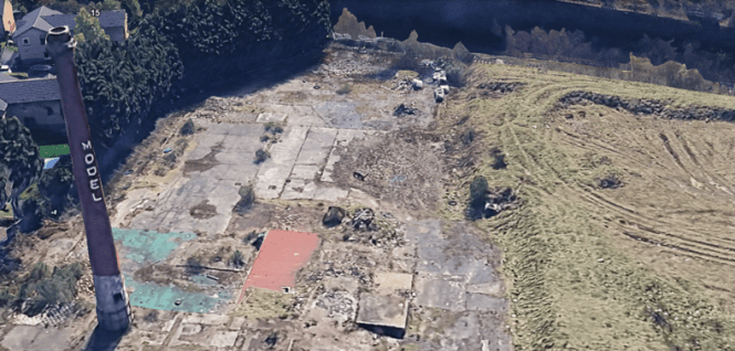 A high-angle 3D satellite image showing the abandoned, demolished site of a former industrial facility. A tall brick chimney stack stands prominently on the left side, with the word "MODEL" painted vertically down its side in white letters. The rest of the plot consists of overgrown concrete foundations, patches of dirt, scattered debris, and vegetation. The site is bordered by thick green trees on the left and an earthen bank or field on the right.